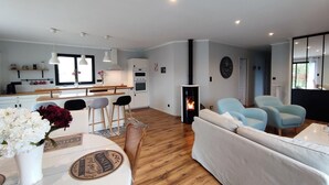 Un salon moderne avec une cheminée, un coin repas avec une table ronde et des chaises, et une cuisine avec des meubles blancs et une fenêtre.