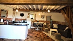 A rustic kitchen with a central island, a dining area with chairs, a sofa, and a clock on the wall. - Gîtes de France® - Le Colombier d'Yville (HERONCHELLES)