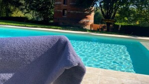 A pool with clear blue water, a brick building in the background, and a purple towel on a chair. - Gîtes de France® - Le Colombier d'Yville (HERONCHELLES)