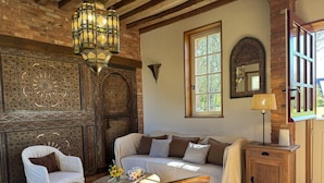 A cozy room with a white sofa, a wicker chair, a wooden cabinet, and a large window with a view of greenery. - Gîtes de France® - La Maison Rouge (BOIS HEROULT)