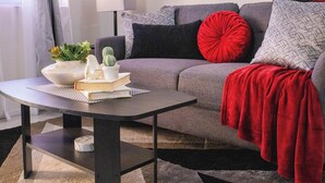 Una sala de estar moderna con un sofá gris, cojines rojos y estampados, una pequeña mesa con un libro, una lámpara y una planta.