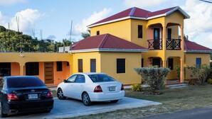 Una casa amarilla de dos pisos, con techo rojo, un balcón y una entrada para coches con dos vehículos estacionados.