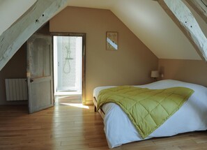 Une chambre avec un plafond en pente, un lit recouvert d’une couverture verte, une porte donnant sur une salle de bain et une petite lampe fixée au mur.