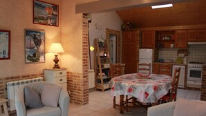 A cozy living room with a dining area, a fireplace, and a kitchen with wooden cabinets and a tiled backsplash. - Gîtes de France® - Auleclairlogis (ETAINHUS)