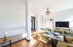 Quarto de hotel moderno com uma cama, área de refeições com mesa e cadeiras, televisão e uma janela grande.