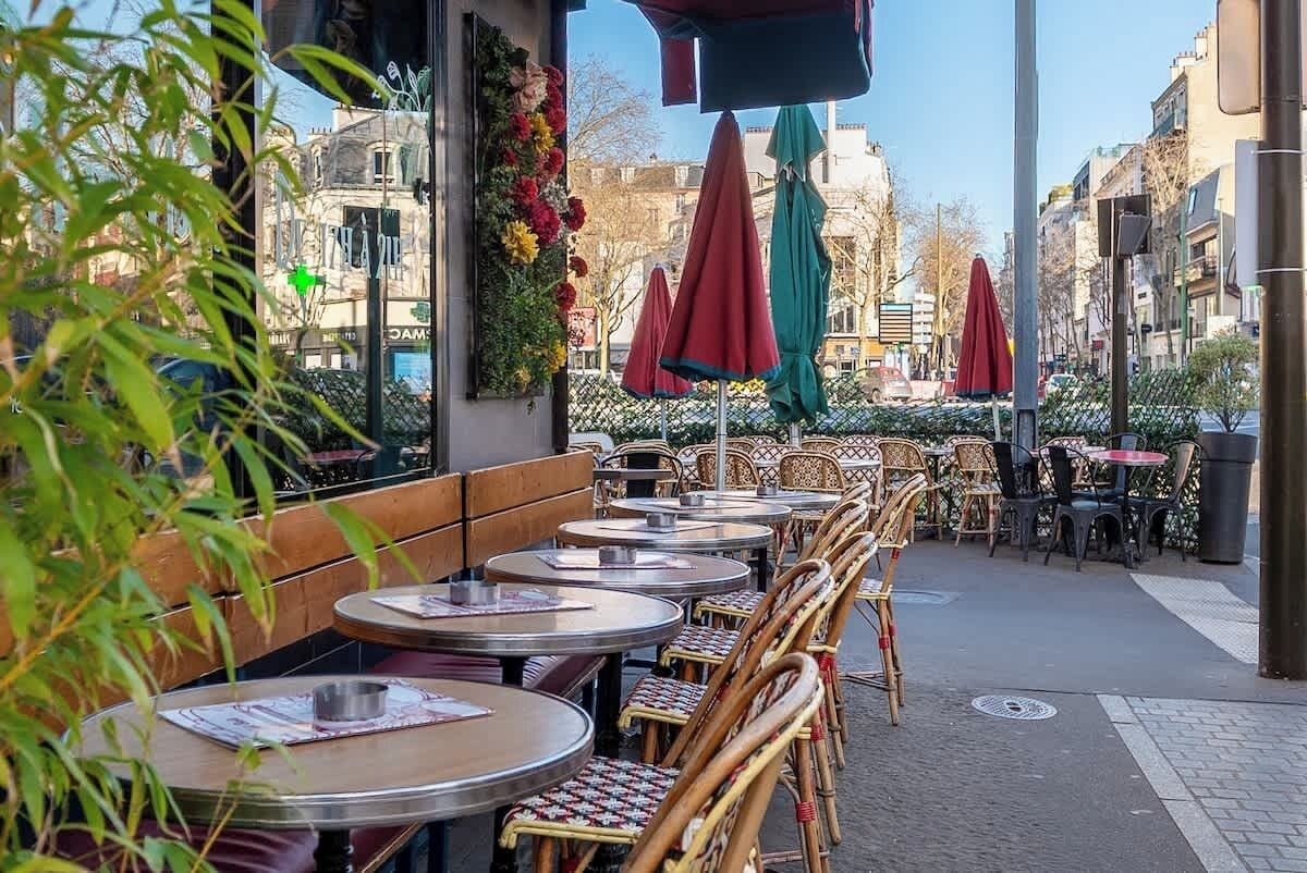 Ein Straßencafé mit runden Tischen, Korbstühlen, einer Bank und Sonnenschirmen.