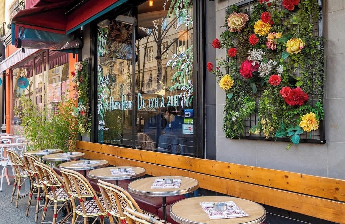 An outdoor cafe with round tables and wicker chairs, a glass window with a floral display, and a sign reading 'Happy Hour.'