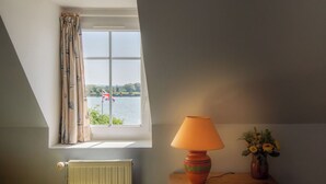 Une pièce avec une fenêtre donnant sur un plan d’eau, un radiateur, une lampe et un vase rempli de fleurs.
