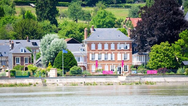 Une ville riveraine dotée de bâtiments historiques, d’une imposante maison en briques aux fenêtres blanches et d’une végétation luxuriante.