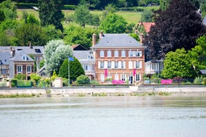 Une ville riveraine dotée de bâtiments historiques, d’une imposante maison en briques aux fenêtres blanches et d’une végétation luxuriante.
