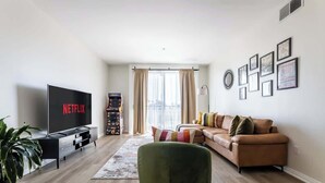 Una sala de estar moderna con una televisión de pantalla plana, un sofá, un reposapiés verde y un ventanal con cortinas.