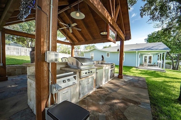 An outdoor kitchen area with a grill, sink, and storage cabinets, under a wooden pergola with a stone floor.