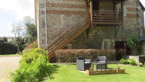 Un bâtiment en pierre avec un escalier en bois menant à un balcon. On y trouve des espaces aménagés pour s’asseoir à l’extérieur et une pelouse bien entretenue.