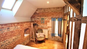 Une chambre à coucher aménagée dans un grenier, avec des murs en briques apparentes, un plancher en bois, un fauteuil suspendu et une lucarne.