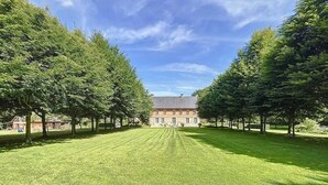 Une vaste étendue herbeuse, à ciel ouvert, où les arbres sont disposés de manière symétrique, et un bâtiment central doté d’un toit en pente.