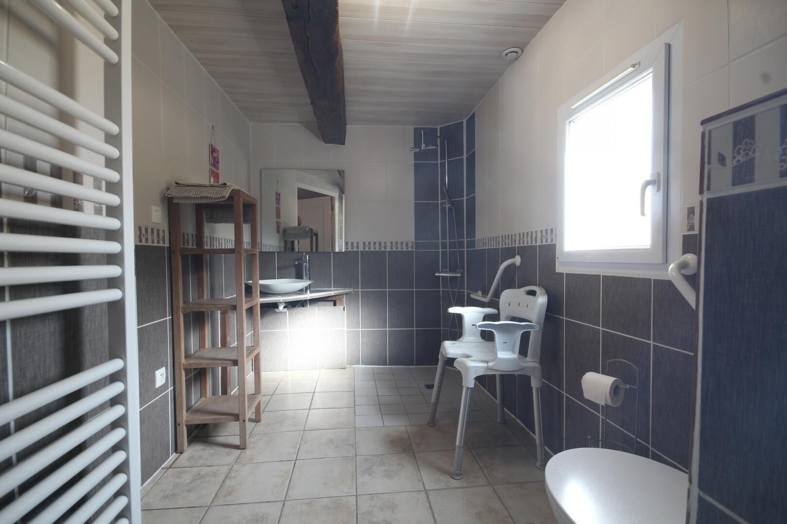 A bathroom with a toilet, sink, and shower. There is a wooden shelf and a window.
