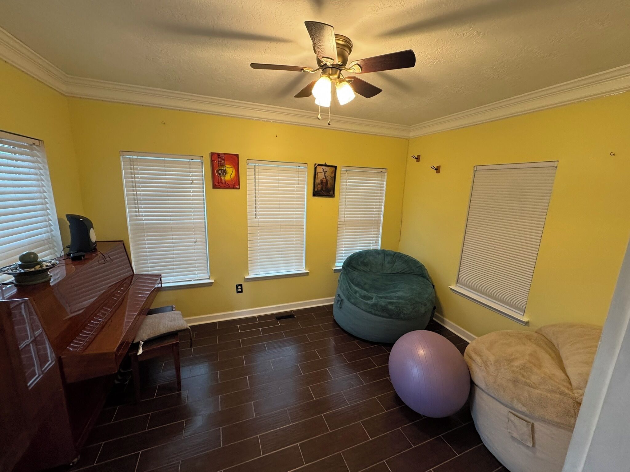 A room with yellow walls, a ceiling fan, a piano, exercise balls, and a bean bag chair.