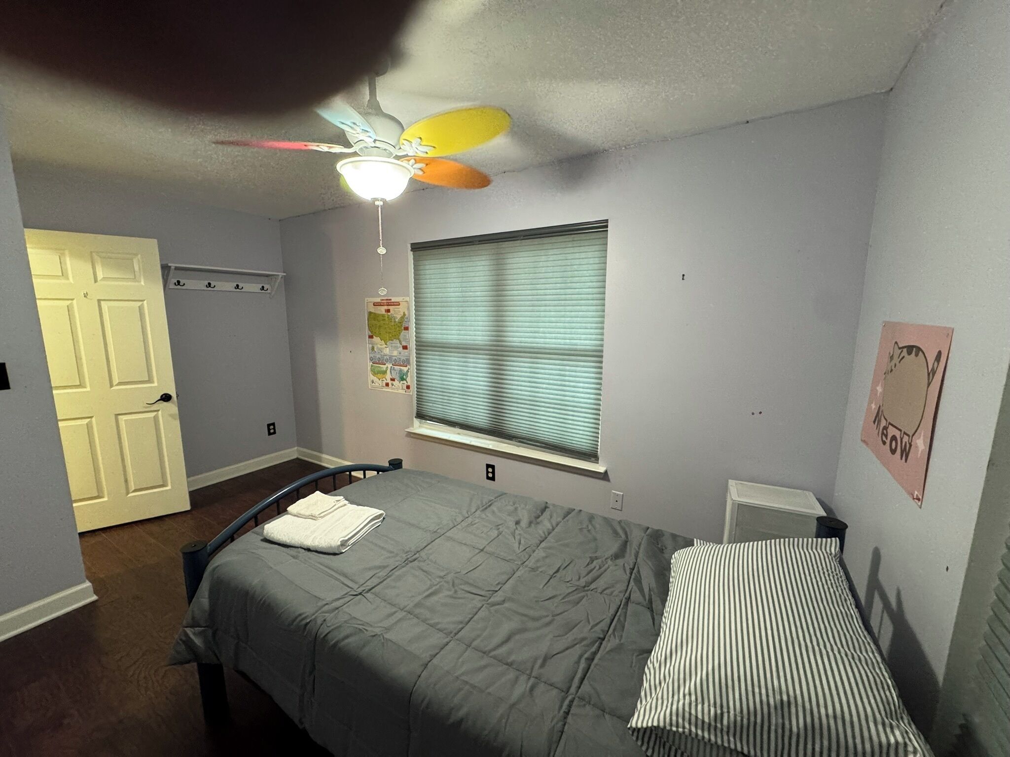 Une chambre à coucher avec un lit, un ventilateur de plafond, une fenêtre avec des stores, une porte et un mur orné d’une carte et d’une affiche.