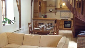 Ein gemütliches Wohnzimmer mit einem beige farbenen Sofa, einem Essbereich mit Holzmöbeln und einer Küche mit Holzausbau und einem Kamin aus Ziegelsteinen.