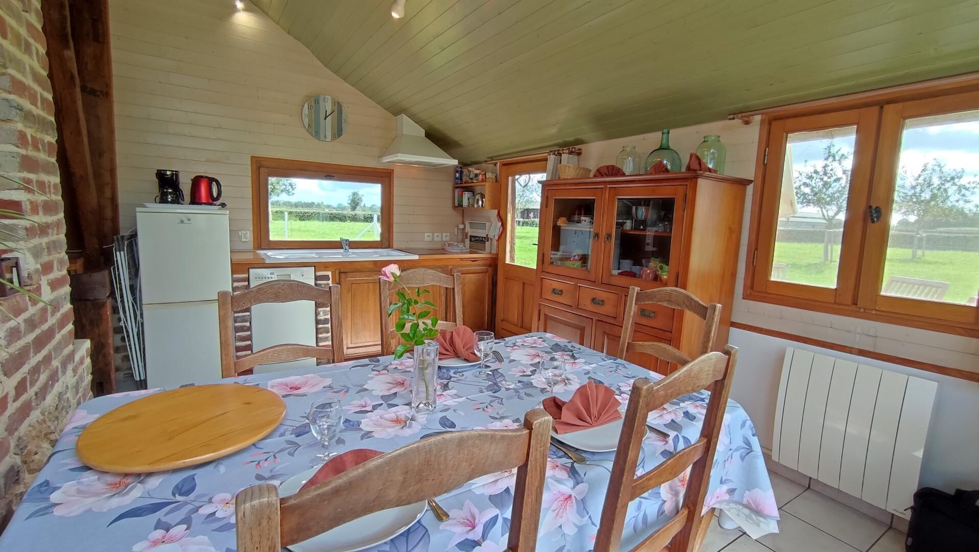 A cozy kitchen with wooden cabinets, a dining table set for two, and a view of the garden through the windows.