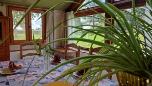 Un coin repas avec une table dressée pour deux personnes, un vase contenant une seule rose et une vue sur un paysage verdoyant par de grandes fenêtres.