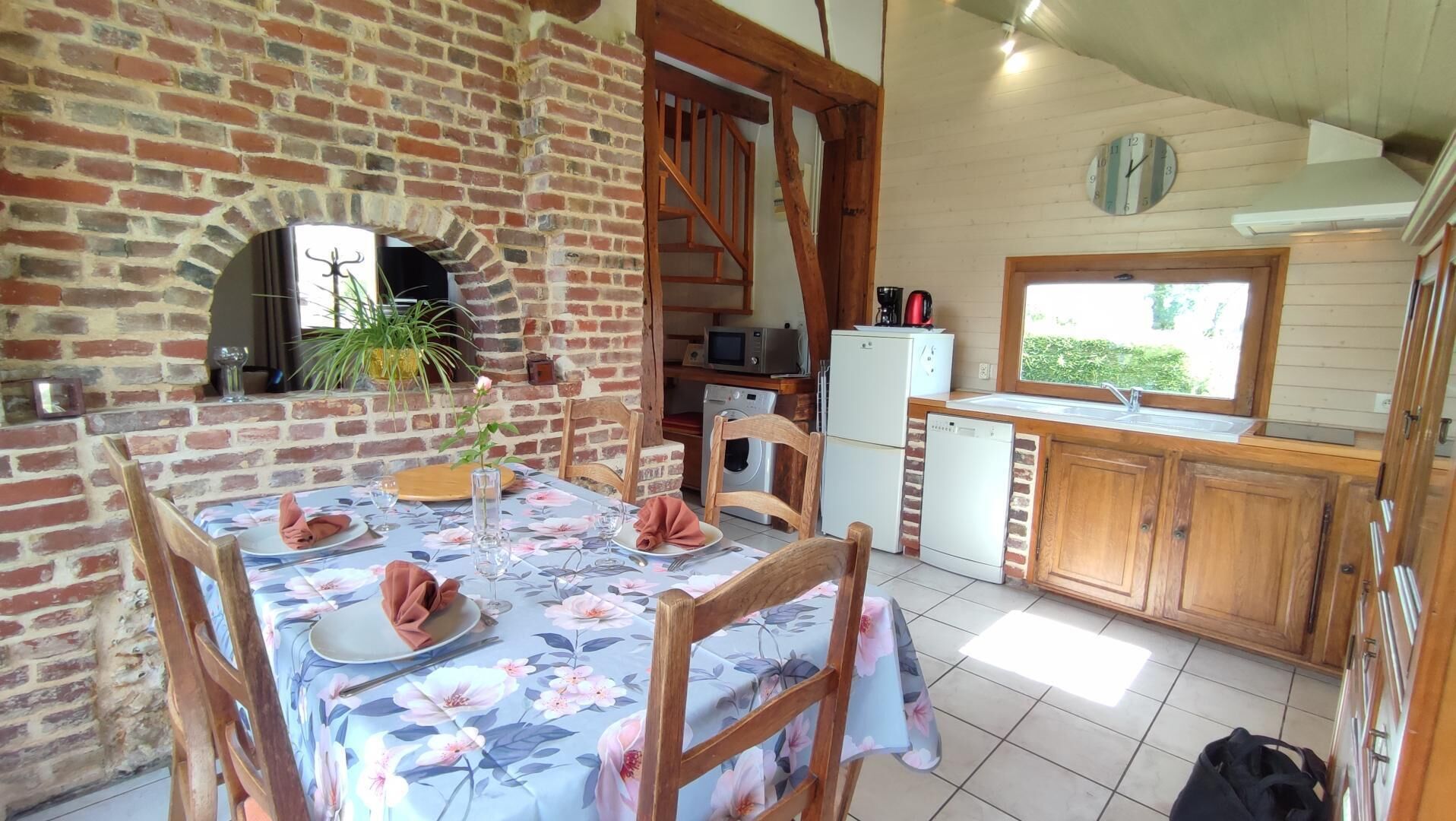A rustic kitchen with a brick wall, wooden cabinets, and a dining area with a floral tablecloth.