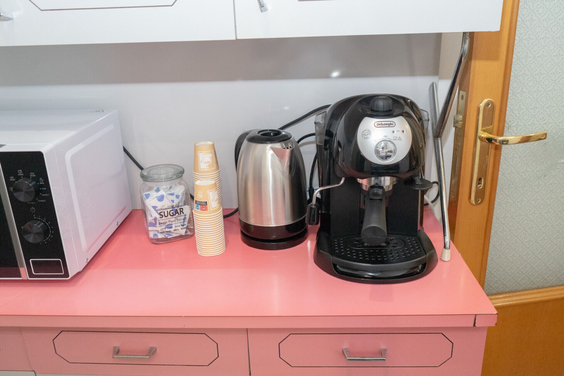 Eine Küchenarbeitsplatte mit Mikrowelle, Kaffeemaschine, Zuckerglas und gestapelten Papierbechern.