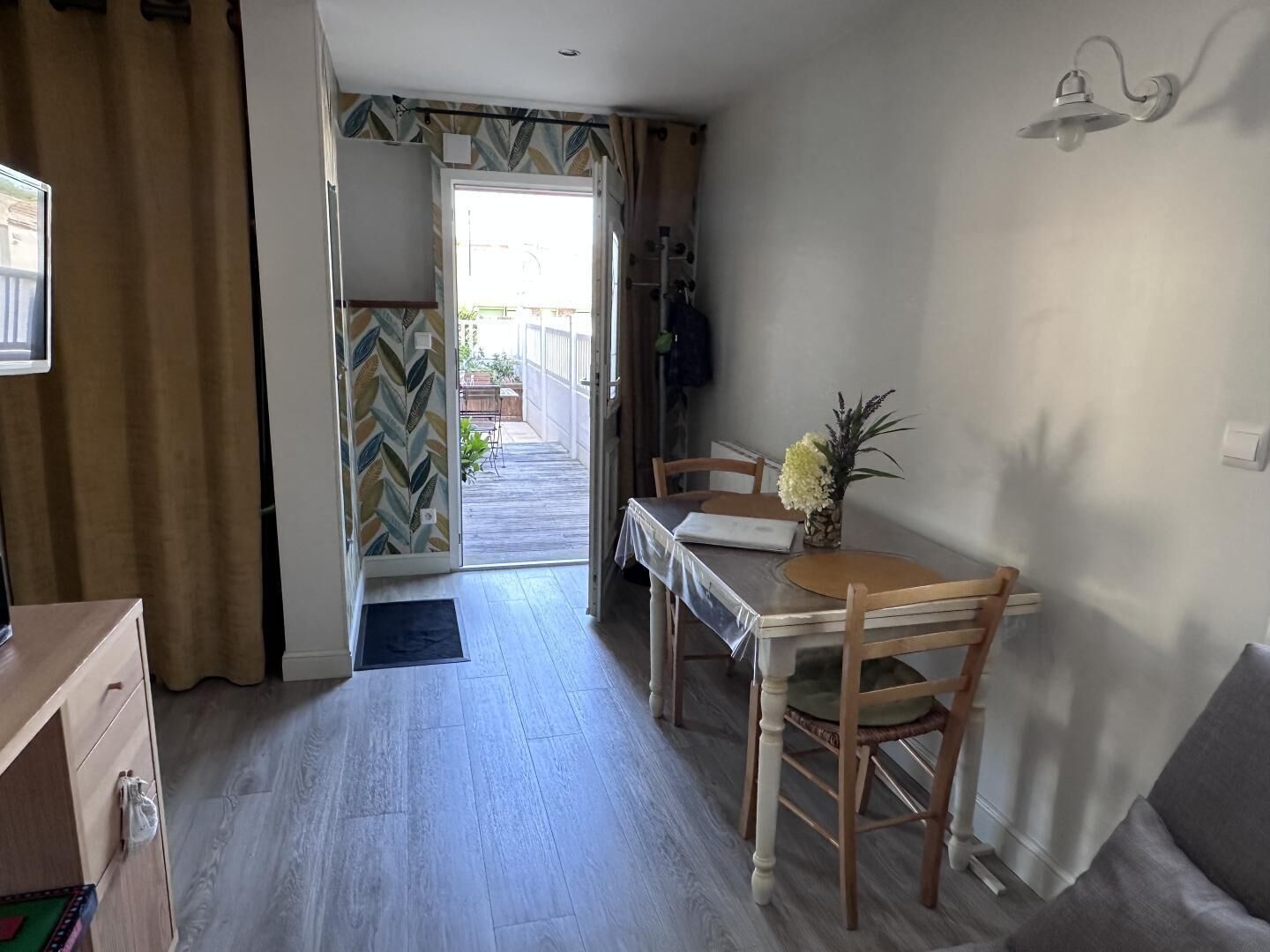 Une pièce comprenant une table à manger en bois, des chaises, un petit vase avec des fleurs, un téléviseur et une porte donnant sur un espace extérieur.