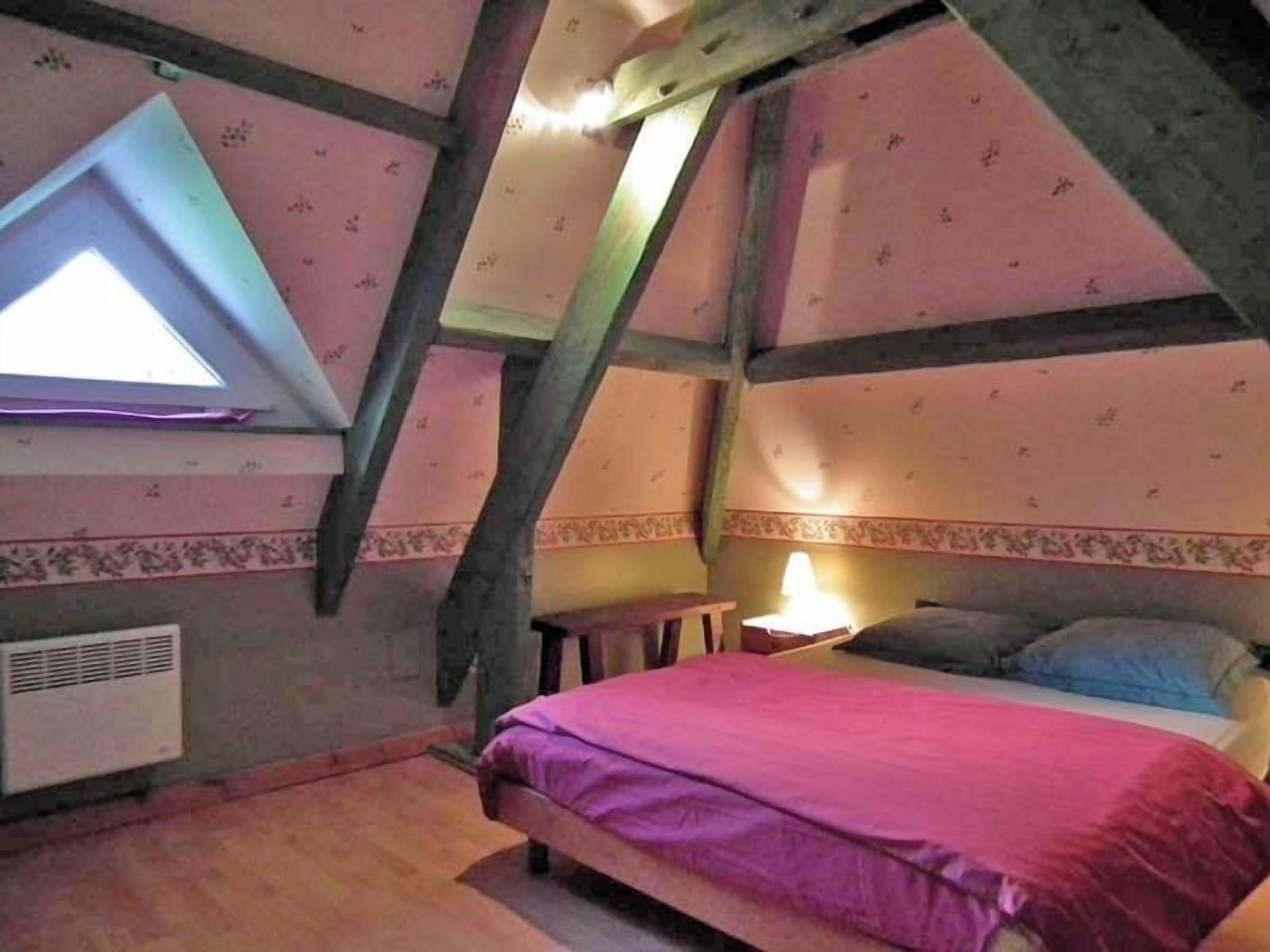 Une chambre avec un plafond en pente, un lit recouvert d’une couverture rose, une petite table de chevet et une lampe.