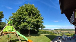 Een speeltuin met een glijbaan en schommels, een grote boom en een terrasgedeelte met stoelen en een tafel.