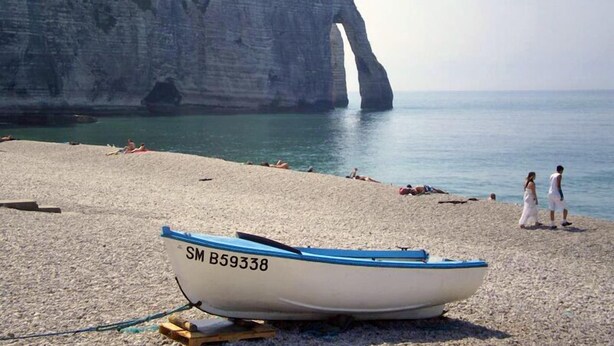 Un barco con la matrícula SM B59338 en una playa pedregosa con gente y un acantilado al fondo.