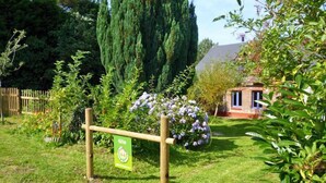 Un jardín con un cartel de madera, abundante vegetación y un pequeño edificio de ladrillo al fondo.
