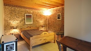 Un dormitorio con una cama de madera, una mesita de noche con lámpara, una pequeña mesa y una silla. La habitación tiene paredes de piedra y techo de madera.