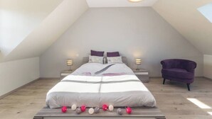 A bedroom with a sloped ceiling, a bed with white bedding, two bedside lamps, and a purple armchair.