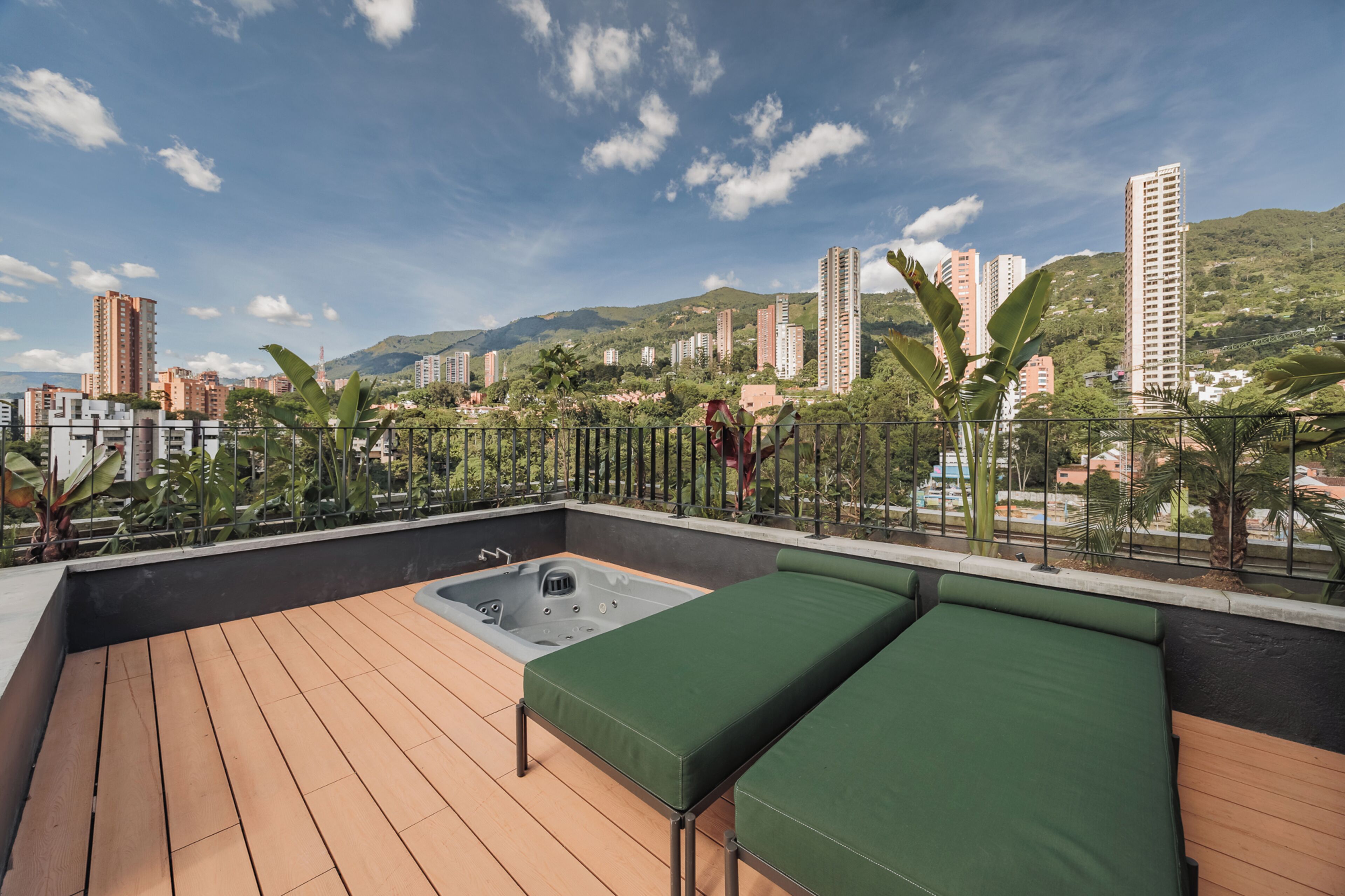 A rooftop terrace with a hot tub, green seating, and a view of a cityscape with buildings and hills.