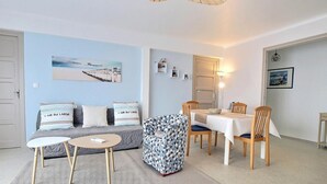 Un salon comprenant un canapé, un fauteuil à motifs, une table ronde et une table à manger avec des chaises. On y trouve également des étagères murales et un tableau.