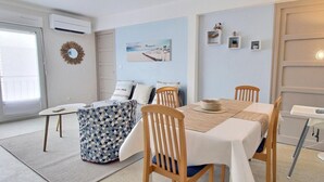 Une pièce moderne et bien éclairée, avec une table à manger dressée pour quatre personnes, entourée de chaises en bois. On aperçoit un pouf à motifs et une petite table d’appoint avec une lampe. La pièce est dotée d’une grande fenêtre, d’un climatiseur mural et d’un miroir décoratif.