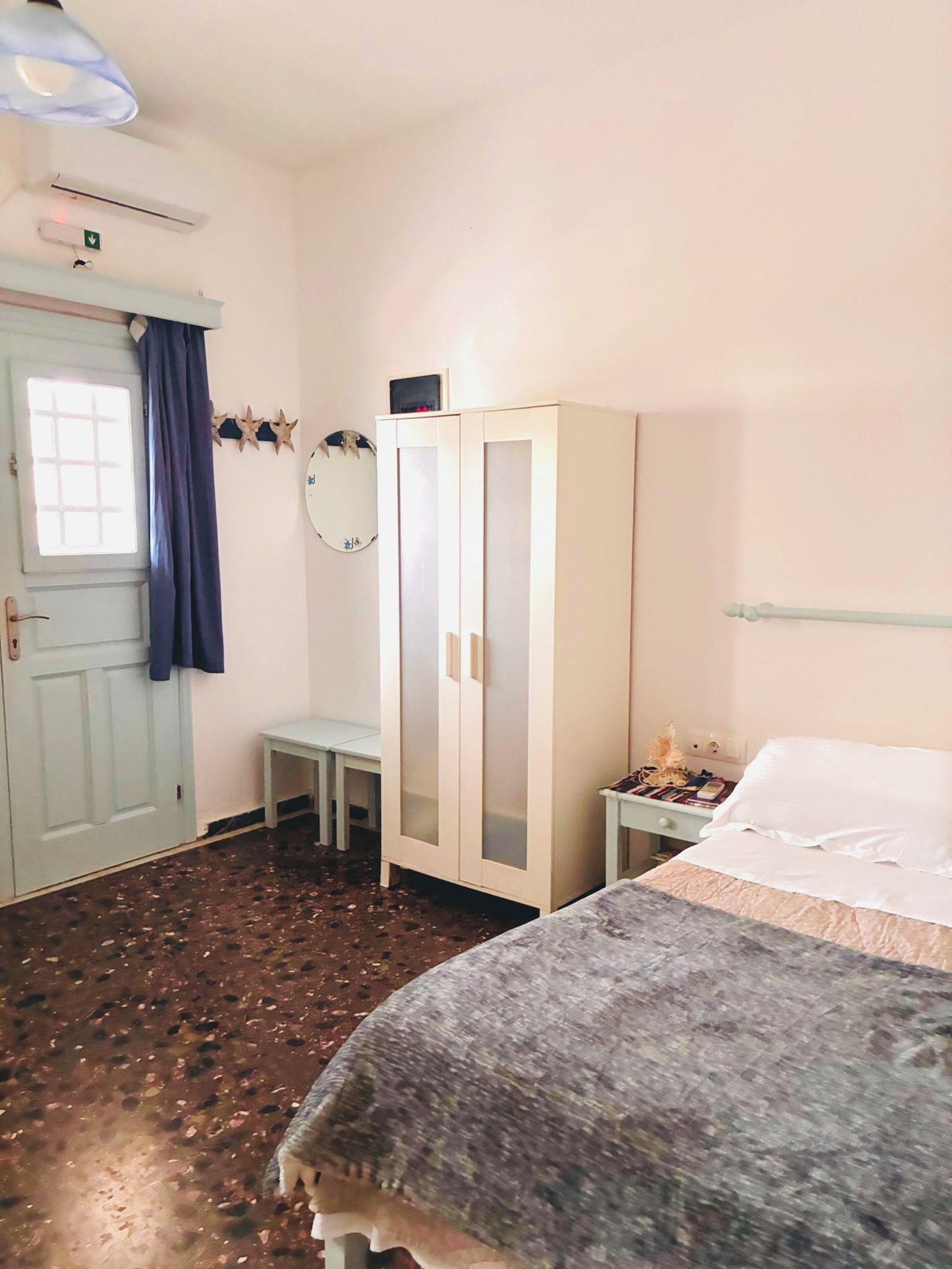A room with a bed, a wardrobe, a small table, a chair, a window with curtains, and a wall-mounted mirror.