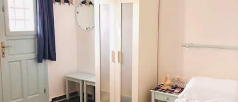 A room with a bed, a wardrobe, a small table, a chair, a window with curtains, and a wall-mounted mirror.