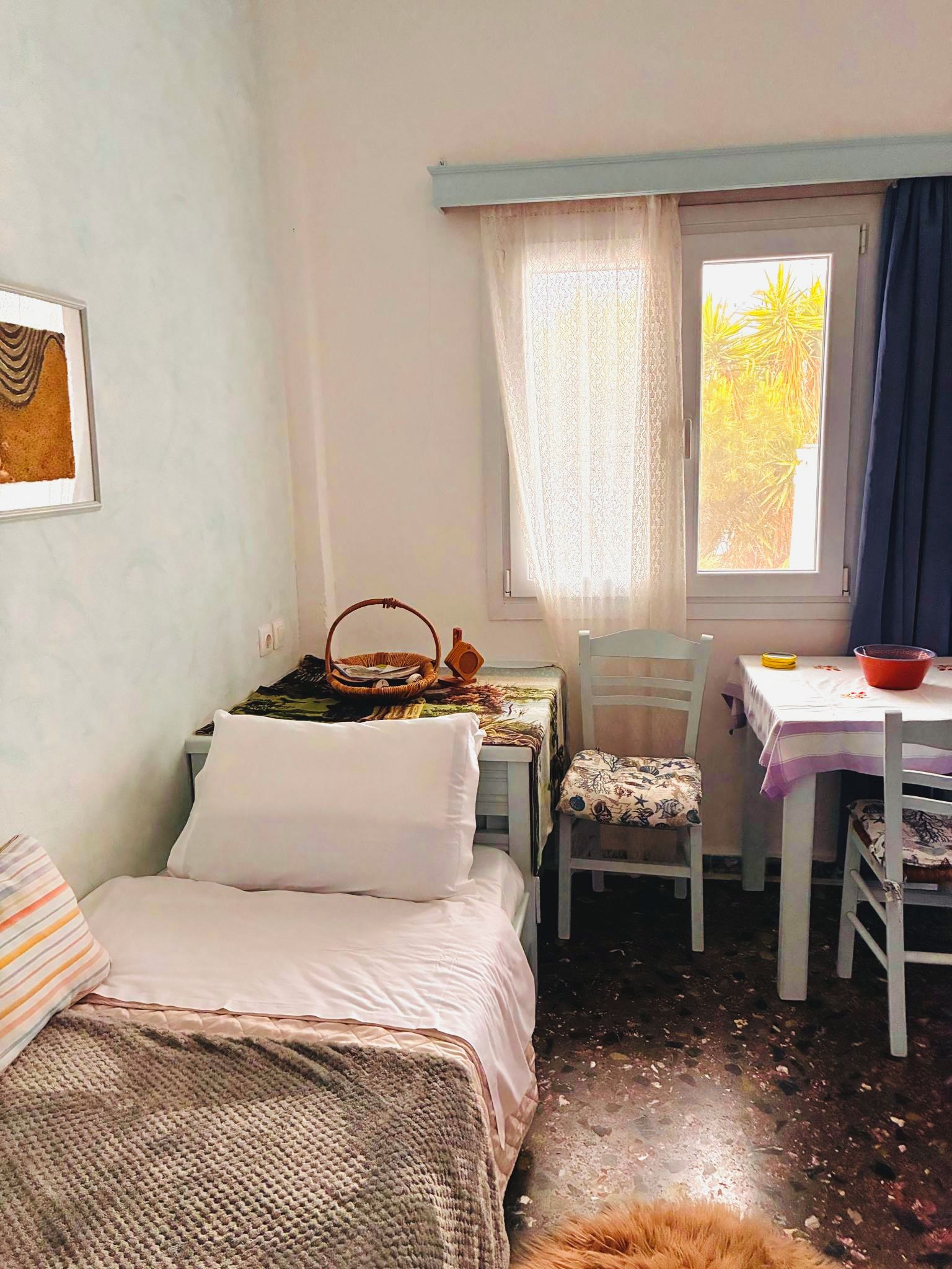 Quarto com cama, uma pequena mesa com cadeira, janela com cortinas e prateleira na parede.