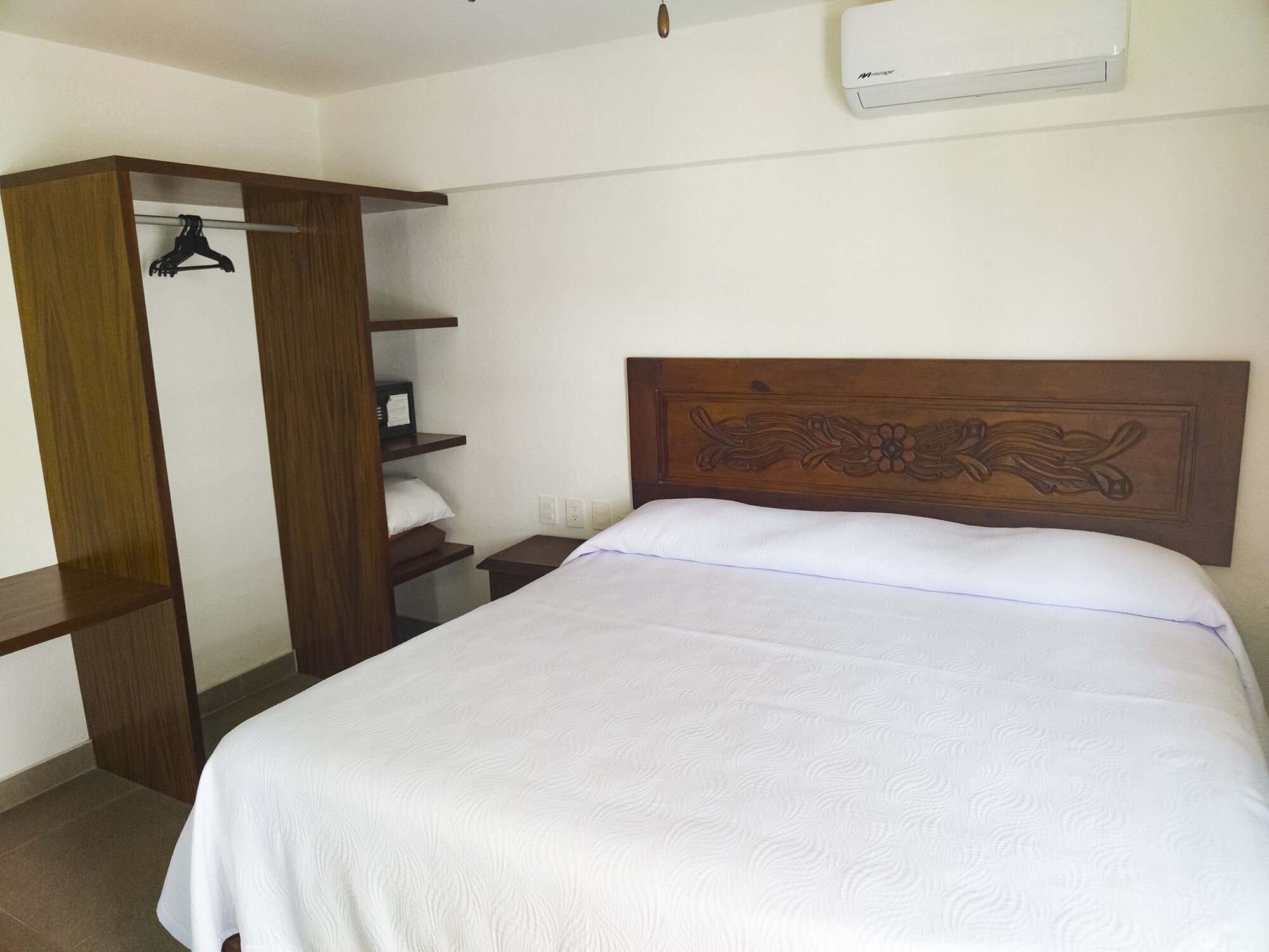 A bedroom with a wooden headboard, a bed with white bedding, a wooden wardrobe, and a wall-mounted air conditioner.