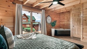 A wooden cabin bedroom with a bed, a clock, a potted plant, and a view of a wooden house outside. - Spring Break Retreat: Gorgeous Views, Sauna & Spa (Sevierville)