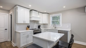 A modern kitchen with white cabinetry, a central island, and a window with blinds. - The Nomad 48-person Hillsboro Village Retreat Studio & 1BR Spaces (Nashville)