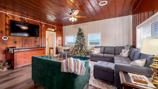 A cozy living room with a wooden ceiling, a flat-screen TV mounted on the wall, a green armchair, a gray sectional sofa, and a Christmas tree. - Incredible Views! Historic Branson Home, two Porch Swings (Branson)