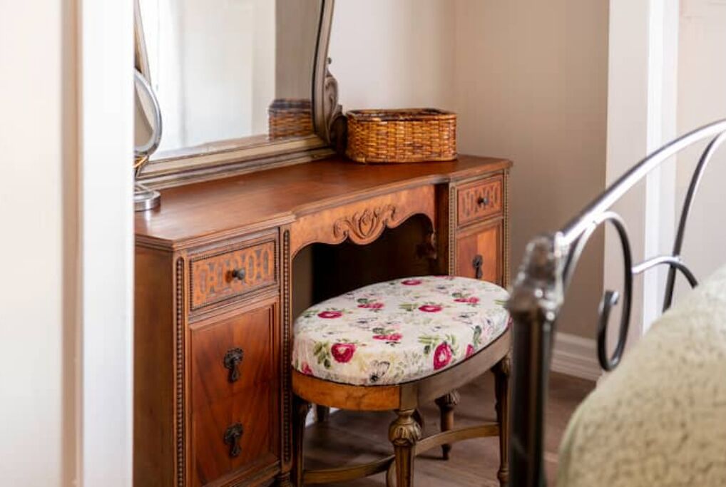 A wooden vanity with a mirror, a wicker basket, and a floral upholstered stool.