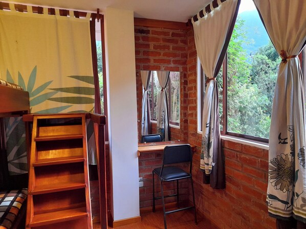 A rustic room with a brick wall, a wooden desk, a chair, and a window with curtains.