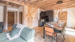 Ein modernes Interieur mit Holzbalkendecke, einem Essbereich mit langem Tisch für sechs Personen und einer Treppe, die zu einer oberen Ebene führt.