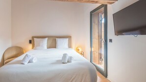 Ein ordentlich bezogenes Bett mit weißen Leinen und zwei aufgerollten Handtüchern, ein Holz-Kopfteil, eine Nachttischlampe und ein an der Wand montierter Fernseher.