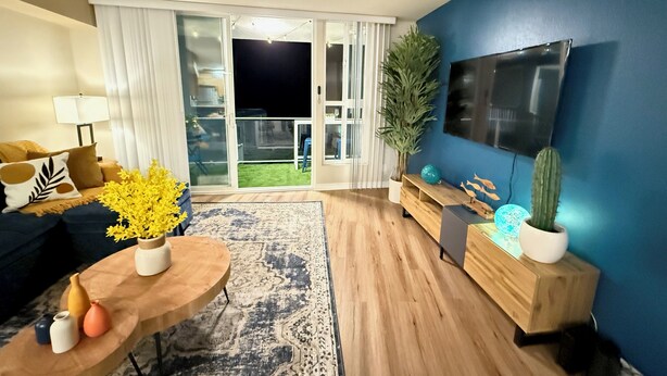 A modern living room with a wooden floor, a TV mounted on the wall, a sofa, a coffee table with vases, and a sliding glass door leading to a balcony. - Oceanfront 1 BR w/ Pool and Heated Pool (Redondo Beach)