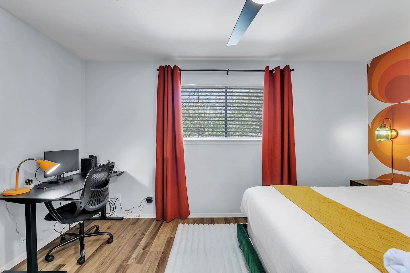 A modern bedroom with a bed, desk, and chair, featuring a window with curtains and a ceiling light.